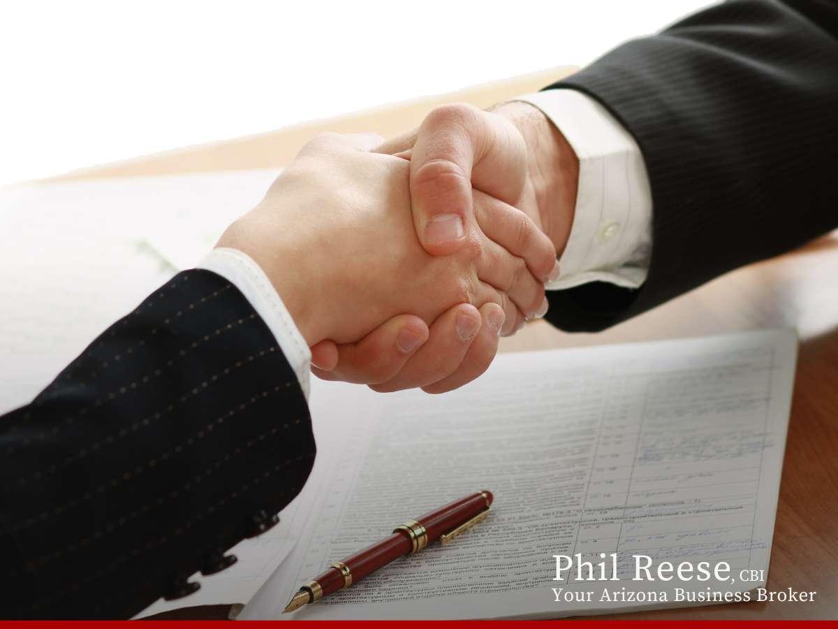 Two professionals shaking hands over a table with financial statements and a pen, signifying a business sale.